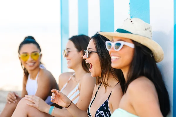 Four young women in bikinis laughing with each other leaning against the beach wall.