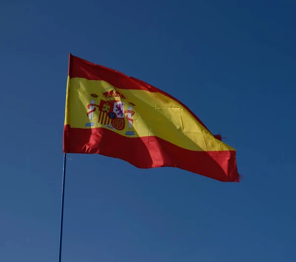 Испанский национальный флаг, размахивающий на фоне голубого неба. Флаг на флагштоке развевается на ветру. Красный с желтым флагом Испании, дующий на ветру против голубого неба — стоковое фото