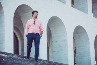 Çizgili gömleği ve pantolonu dışarıda elleri ceplerinde genç bir adam. Merdivenleri çıkarken arkasında bir bina vardı. Gömlekte güneş gözlüğü var..