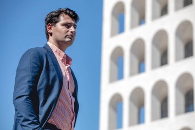 Bir binanın önünde duran bir iş adamı. Tişört ve takım elbise giyiyordu. Eller cebe..