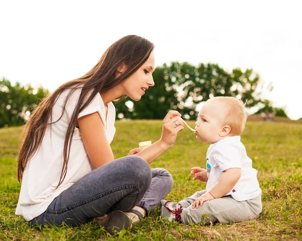 Young beautiful mother feeding her baby puree outdoors in sunlight — Stock Photo, Image