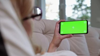 Kanepede uzanan ve internet üzerinden krom tuşlu akıllı telefondan video izleyen bir kadın.