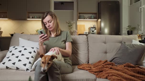Жінка сидить на дивані з собакою і використовує смартфон — стокове відео