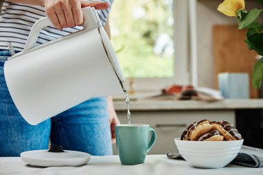 Tea time, Woman pouring boiled water from electric kettle into cup for preparing tea, Morning breakfast at kitchen