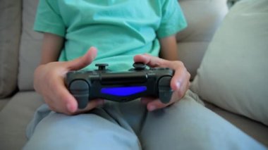Çocuk oturma odasında kanepede oturuyor ve elinde oyun konsoluyla video oyunu oynuyor. Çevrimiçi eğlence ve eğlence aktivitesi.