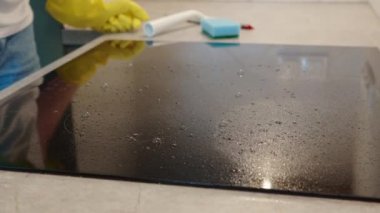 Woman in yellow rubber gloves cleans kitchen induction hob with cleaning spray and sponge. Clear kitchen appliance, Daily chores and housework