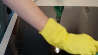 Woman in yellow rubber gloves cleans kitchen induction hob with cleaning spray and sponge. Clear kitchen appliance, Daily chores and housework