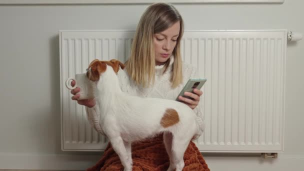 ヨーロッパの家庭では寒い冬 ガスと電気の上昇コスト 生活室での女性と犬の凍結 暖房ラジエーターの近くの毛布の下で暖まる — ストック動画