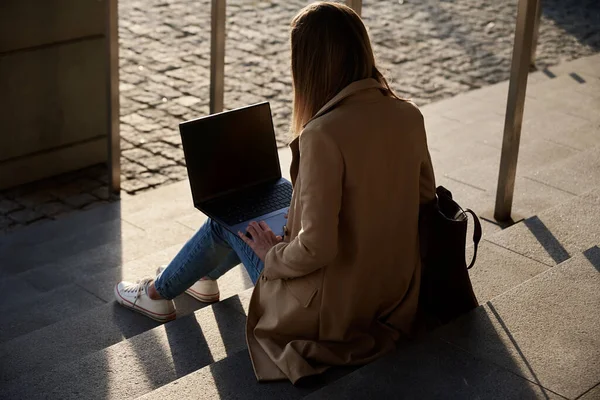 Woman works remotely at city street, use laptop and sitting on stairs. Female freelancer working online at summer day. Remote work and freelance