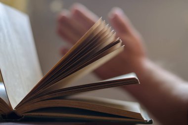 Eski açık bir kitap ve iki adamın elinin odaklanmış görüntüsü. Arka planda dua etmek için avuç içleri katlanmış. Kapak kitabının tozlu sayfaları. Yan görüş. İçeride. Seçici odaklanma. Bulanık, gürültü, tahıl etkisi.