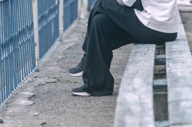 Kadının alt kısmı bankta oturuyor. Geniş siyah pantolonlu, beyaz gömlekli ve spor ayakkabılı bir kadın. Yan görüş. Seçici odak.