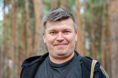 Uzun çam ağaçlarının önündeki orta yaşlı bir adamın portresi. Kısa saçlı ve gri saçlı yetişkin bir adam. Kahverengi gözlü kişi kameraya bakıyor. Seçici odak.