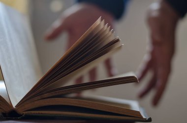 Eski açık bir kitabın ve iki açık erkek elinin odaksız çekimi. Avuçlar arka planda. Kapaklı kitabın sayfaları. Yan görüş. Odanın içinde. Seçici odaklanma. Bulanık, gürültü, tahıl etkisi.