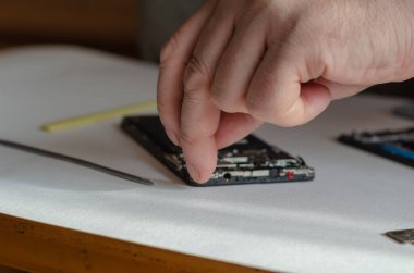 Akıllı telefondaki devre kartının bağlantısını kesen bir adamın yakın çekimi. Siyah parçalanmış telefon açık renkli bir yüzeyde yatıyor. Tamir servisleri. Seçici odak.
