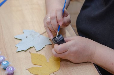 Sanatçı kurumuş yapraklarla resim çizer. Bir kadının elleri kavak yaprağının üzerine siyah guaj boyasıyla fırçalar. Masada akrilik ve guaj var. İçeride. Seçici odak.