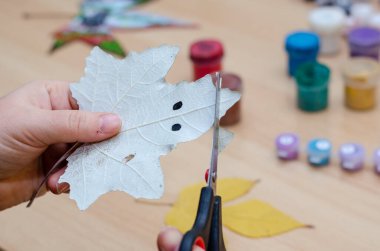 Bir kadın bir kavak yaprağını makasla kesiyor. Üzerine hayalet resmi çizilmiş düşmüş bir yaprak. Arka planda akrilik ve guaj boyaları, fırçalar ve sonbahar yaprakları var. Hobi, yetenek, sanat.