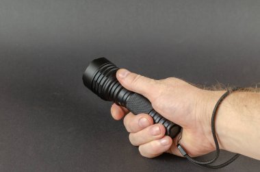 Bir adam siyah bir LED el feneri tutuyor. El askısı gri arka planın sağ tarafına takılmış. Seçici odak.