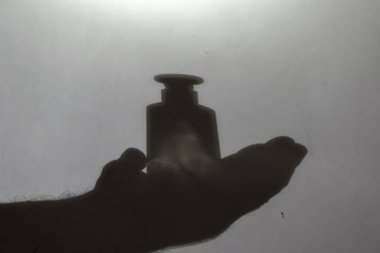 Görüntü, bulanıklık, gürültü, tahıl etkisi. Cam şişeyi tutan siyah bir elin silüeti. Açık gri bir duvarda cam ilaç şişesi ya da parfüm tutan bir adamın çizimi. Seçici odak.
