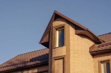 Bulutsuz bir gökyüzüne karşı güzel, modern taştan bir malikanenin tepesinde. Ev sarı fayanslarla kaplı. Kahverengi çatılı ve kar korumalı. Modern gayrimenkul, mimari.