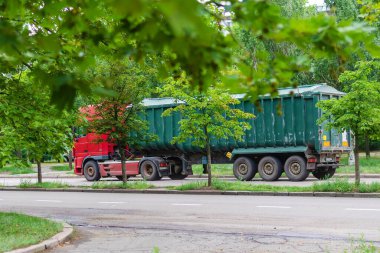 Asfaltlı yolda yeşil römorklu kırmızı traktör. Kestane ve çınar ağaçlarının yeşil yaprakları arasında bir tahıl arabası. Lojistik, endüstri. Nikolaev, Ukrayna - 07 13 2021