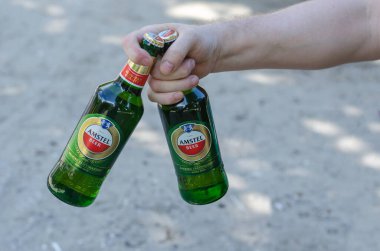 Bir el, kuma karşı iki şişe Amstel birası tutuyor. Yeşil şişe alkollü yetişkin bir erkek. Nikolaev, Ukrayna - 07 13 2021