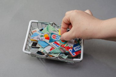 Bir kadının eli, alışveriş arabasından banka kartının bir parçasını çıkarır. Çeşitli bankalardan kesilmiş kredi kartlarıyla dolu metal alışveriş arabası gri bir arka planda. Kiev, Ukrayna - 01 05 2022.