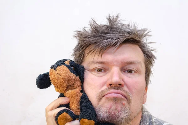 Portrait of a wriggling man with a dog toy. A male with ruffled, tangled hair presses his cheek against the stuffed toy. Indoors. Selective focus.