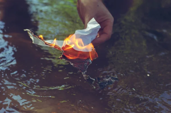 成熟した男は燃える紙を手に持っている 川や川の表面に手で燃える白い紙を砕いた 灰は流れる水の中に落ちる 接近中だ 選択的焦点 — ストック写真