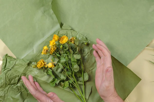 Two men's hands embrace wilting flowers. A bouquet of dying yellow roses against a green background. Selective focus.