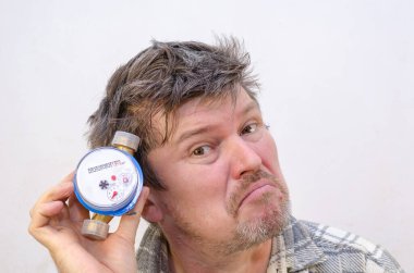 The male grimaces and listens to the water meter with it to his ear. A man with tangled, disheveled hair in front of a light gray wall. Indoors. Selective Focus. clipart