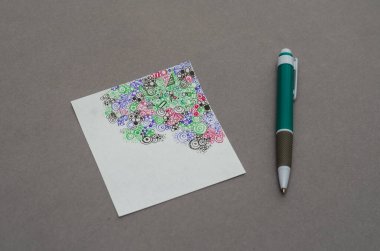 Beyaz kare bir kağıt üzerinde soyut bir çizim ve gri bir yüzey üzerinde duran bir kalem. Farklı geometrik şekillerde çok renkli desenler. Yakın plan. Seçici odak.