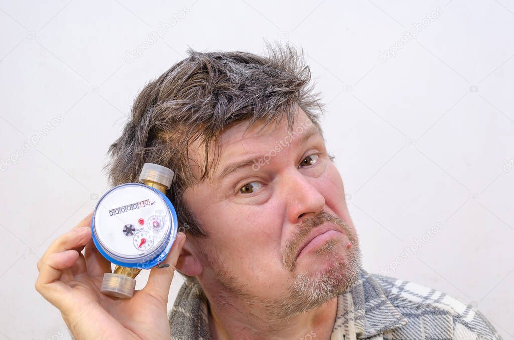 The male grimaces and listens to the water meter with it to his ear. A man with tangled, disheveled hair in front of a light gray wall. Indoors. Selective Focus.