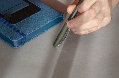 Orta yaşlı adamın elleri arka planda kalemi tutuyor. Kapalı mavi iş günlüğü. Yakın plan. İçeride. Seçici odak.