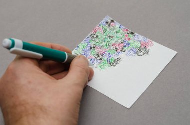 Bir adamın eli beyaz kare bir kağıt üzerine bir kalemle soyut bir çizim yapar. Farklı geometrik şekillerde çok renkli bir desen. Yakın plan. Seçici odak.