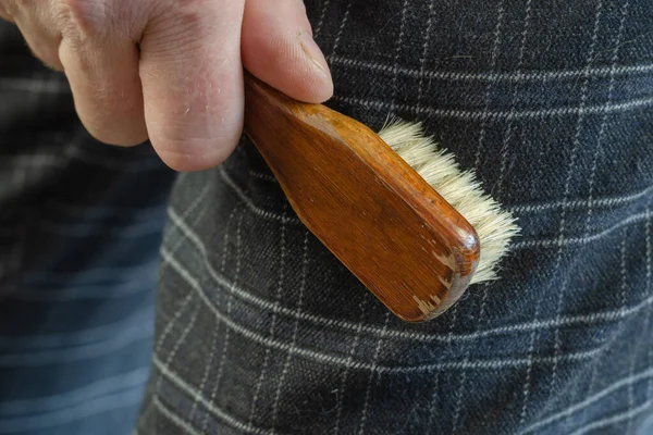 A mature man cleans his pants with a brush. The hand holds a small wooden clothes brush. Close-up. Selective focus. Indoor.