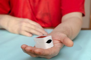 Olgun bir adamın avuç içi beyaz bir nabız oksimetresiyle kameraya uzanır. Arka planda kırmızı tişörtlü bir adam oturuyor. Odanın içinde. Seçici odak.