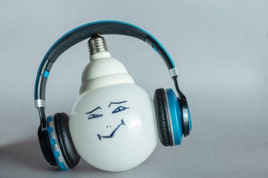 Boyalı yüzü ve kulaklığı olan büyük beyaz bir lamba. Neşeli bir yüzle lambaya kablosuz kulaklık takıldı. Gri arkaplandaki kompozisyon