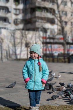 Mavi ceketli bir kız şehir güvercin sürüsünün önünde poz veriyor. Uzun at kuyruklu, beş yaşında bir çocuk. Arka planda kuş yeminin bulanık görüntüsü. Gündüz vakti. İlkbahar.
