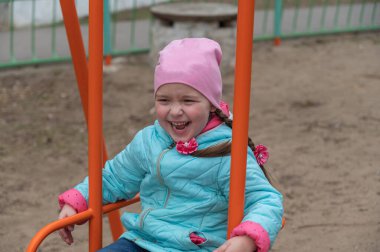 Mavi ceketli bir çocuk salıncakta sallanıyor. At kuyruklu mutlu beş yaşındaki kız çocuk parkında eğleniyor.