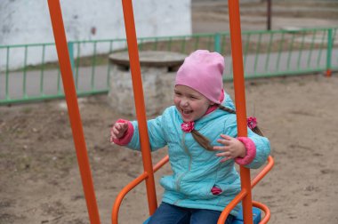 Mavi ceketli bir çocuk salıncakta sallanıyor. At kuyruklu mutlu beş yaşındaki kız çocuk parkında eğleniyor.
