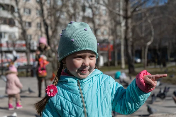 Anaokulu çağında mavi ceketli bir kız işaret parmağıyla sokakta jest yapıyor. Uzun at kuyruklu beş yaşında bir çocuğun portresi. Güneşli bir bahar günü. Yakın plan. Seçici odak.
