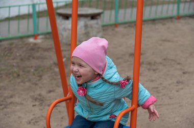 Mavi ceketli bir çocuk salıncakta sallanıyor. At kuyruklu mutlu beş yaşındaki kız çocuk parkında eğleniyor.