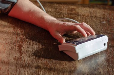 Bir adamın eli dijital tansiyon monitörünün düğmesine basar. Kahverengi ahşap bir masada kan basıncını ölçmek için tıbbi bir cihaz. Odanın içinde. Seçici odak.