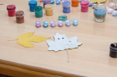 Düşmüş ağaç yaprakları ve bej bir masada çok renkli boya. Kül ve kavak yaprağı. Kavak yaprağına iki siyah nokta çizilir..