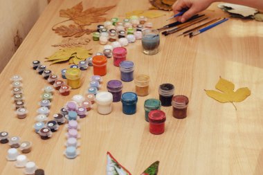 Kurumuş ağaç yaprakları ve bej bir masa üzerindeki boya seti. Bir kadının eli masanın üzerinde duran fırçaları alır. Renkli boyalar..