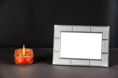 Boş bir yatay fotoğraf çerçevesi ve koyu bir arkaplanda yanan bir mum. Dikdörtgen gümüş fotoğraf çerçevesi ve yuvarlak cam şeffaf bir kavanozda kırmızı mum..