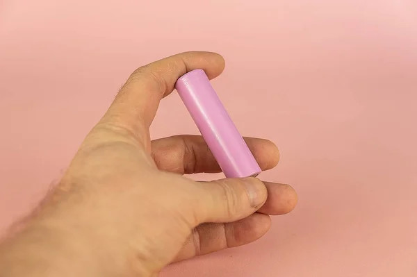 An adult male hand holding a lithium-ion battery against a pink background. The cylindrical rechargeable battery has a high energy density, no memory effect and low self-discharge.