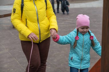 Anne, kızının çocuk kaydırağına tırmanmasına yardım ediyor. Sarı ceketli yetişkin bir kadın köpeği için pembe bir tasma tutuyor. Uzun örgülü bir kız..