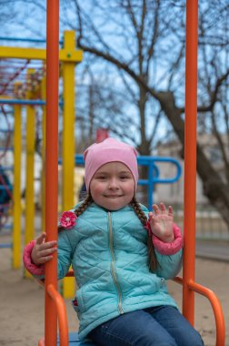 Bir kız salıncakta sallanıyor ve selamlamak için sol elini sallıyor. Çocuk parkında oynayan bir çocuk. Bahar geldi. Seçici Odaklanma.