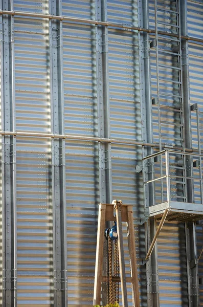 Industrial construction site. Construction of metal cylindrical silos for grain storage. Mechanical cargo winch in front of the gray grain silo